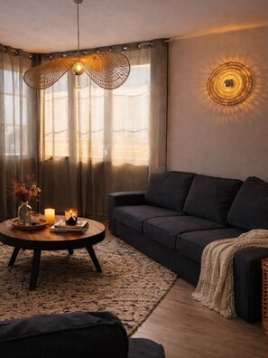A cozy living room with a dark sofa, a round coffee table with candles, a woven light fixture, and a wall-mounted lamp. - New Listing (Royan)