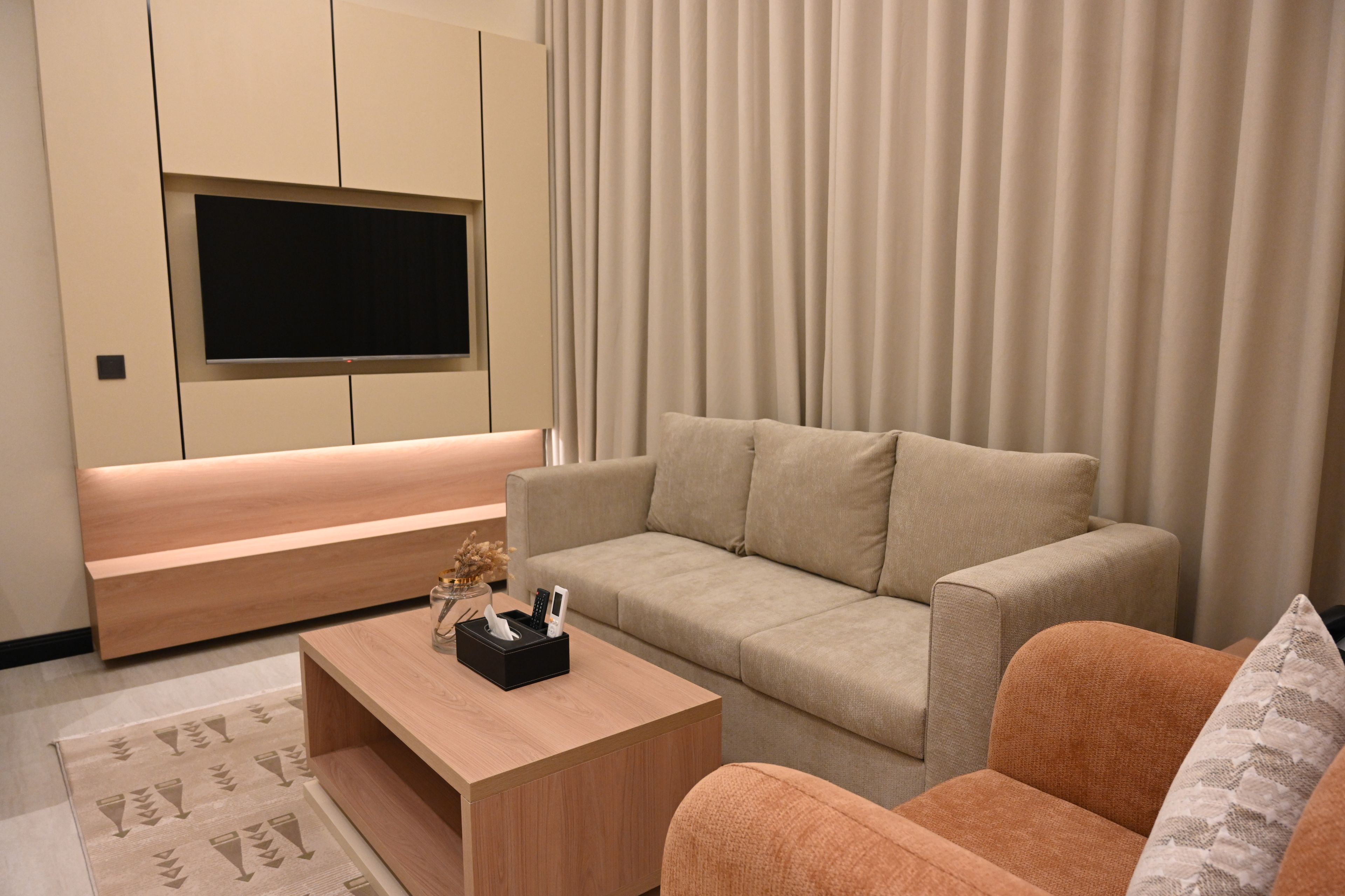 A modern living room with a flat-screen TV mounted on the wall, a beige sofa, a wooden coffee table, and a patterned rug.
