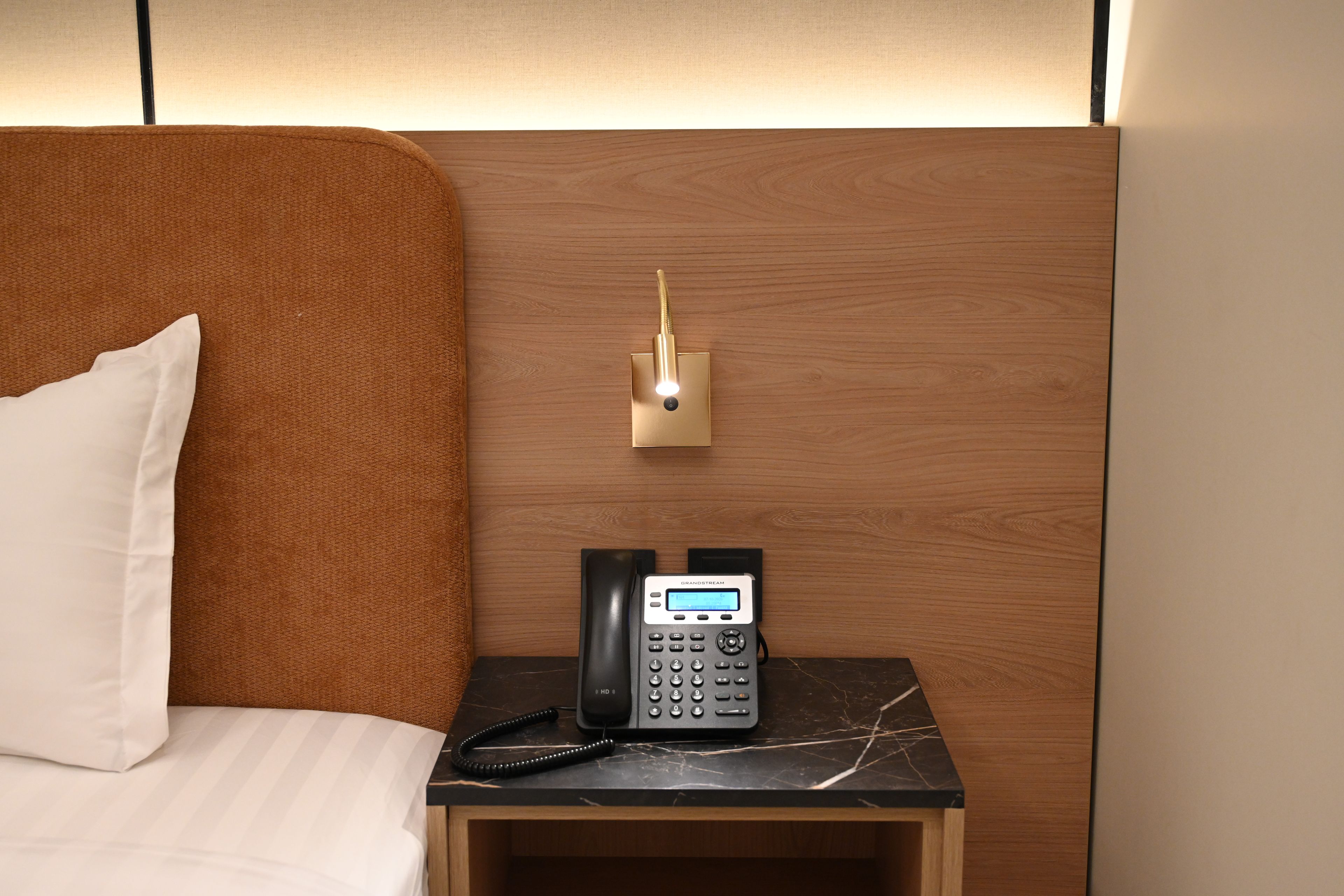 A hotel room with a wooden headboard, a bedside table with a black telephone, and a wall-mounted light fixture.