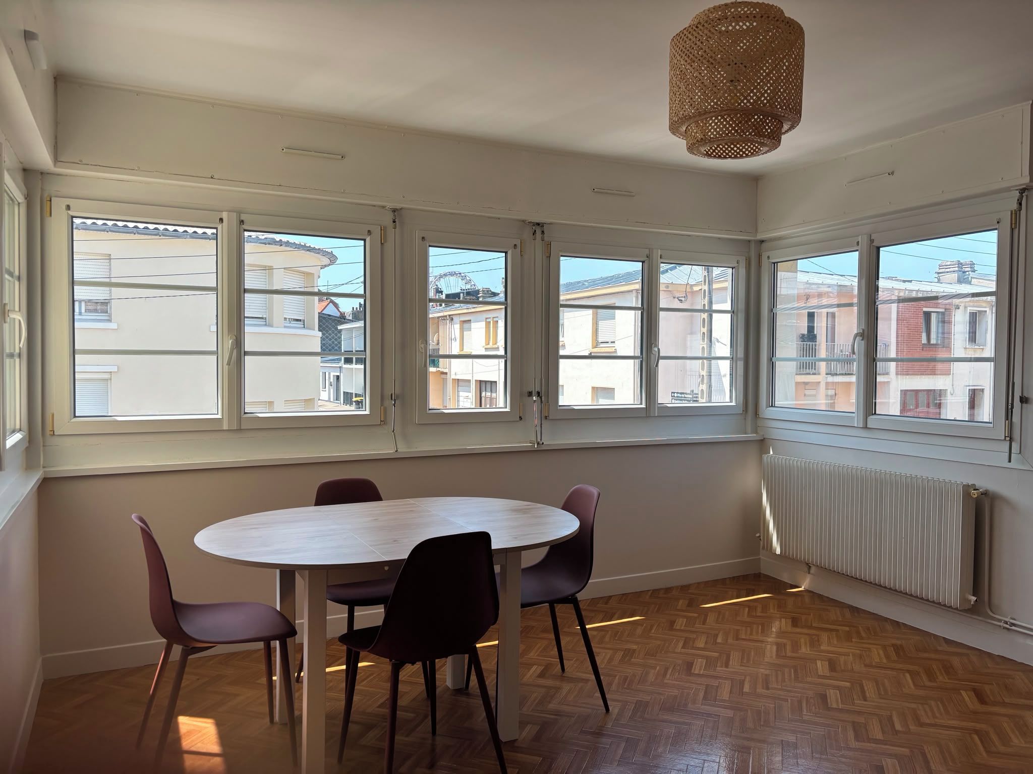 Une pièce avec une table ronde et quatre chaises, de grandes fenêtres donnant sur des immeubles, un radiateur et un luminaire suspendu.