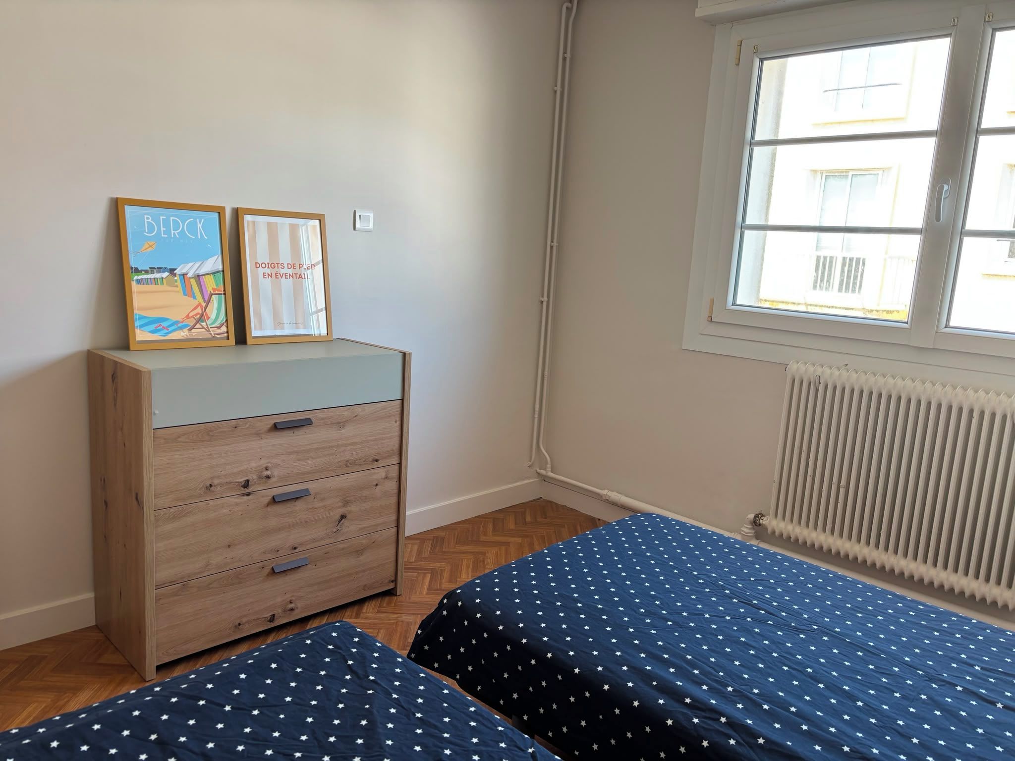Une chambre avec deux lits, une commode en bois et une fenêtre avec des stores.