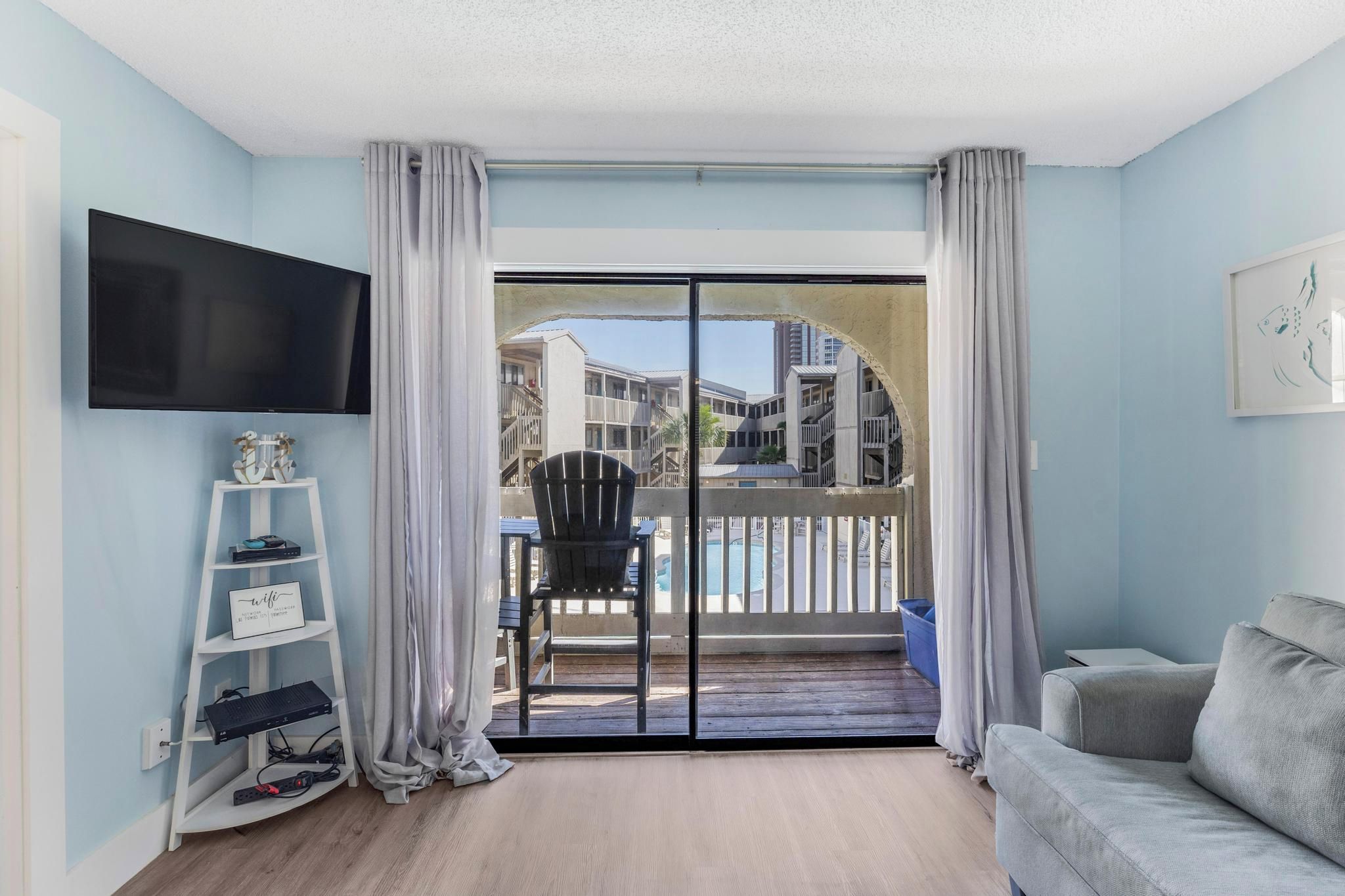A living room with a TV, a sofa, a chair, a small table, and a balcony with a view of buildings.