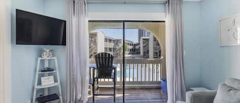 A living room with a TV, a sofa, a chair, a small table, and a balcony with a view of buildings.