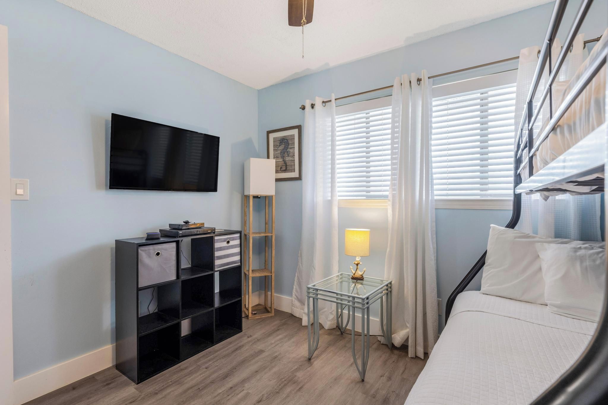 Una habitación con una cama, un televisor montado en la pared, una mesita con una lámpara y un estante con cajas de almacenamiento.