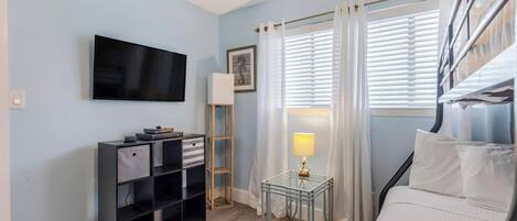 Una habitación con una cama, un televisor montado en la pared, una mesita con una lámpara y un estante con cajas de almacenamiento.