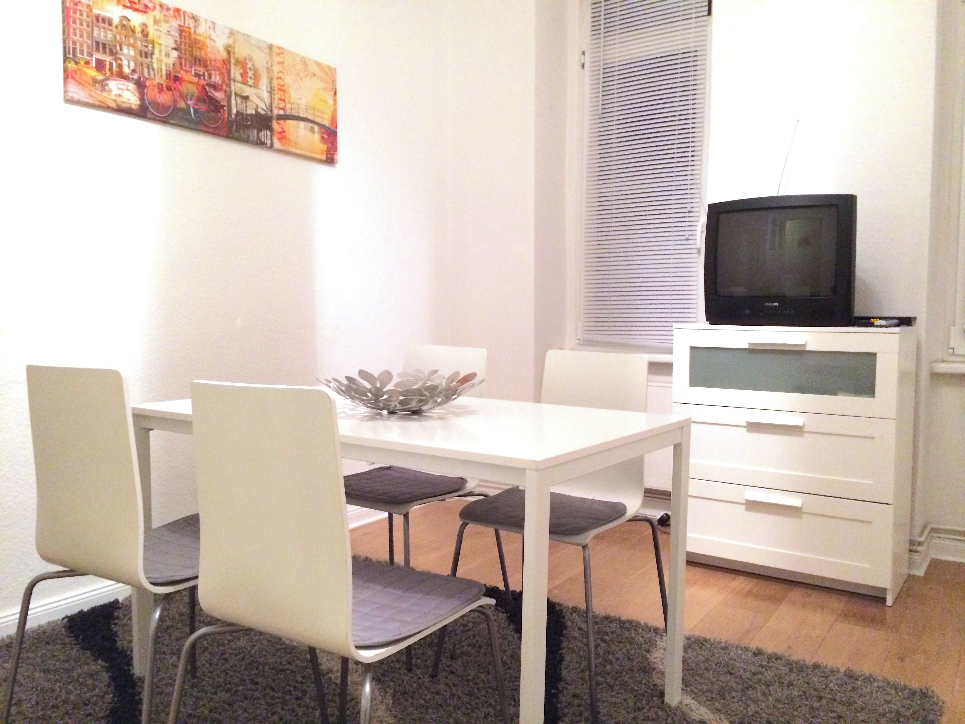 En matplats med ett vitt bord, fyra stolar, en dekorativ skål, en tv på ett skåp och ett fönster med persienner.