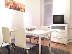 En matplats med ett vitt bord, fyra stolar, en dekorativ skål, en tv på ett skåp och ett fönster med persienner.