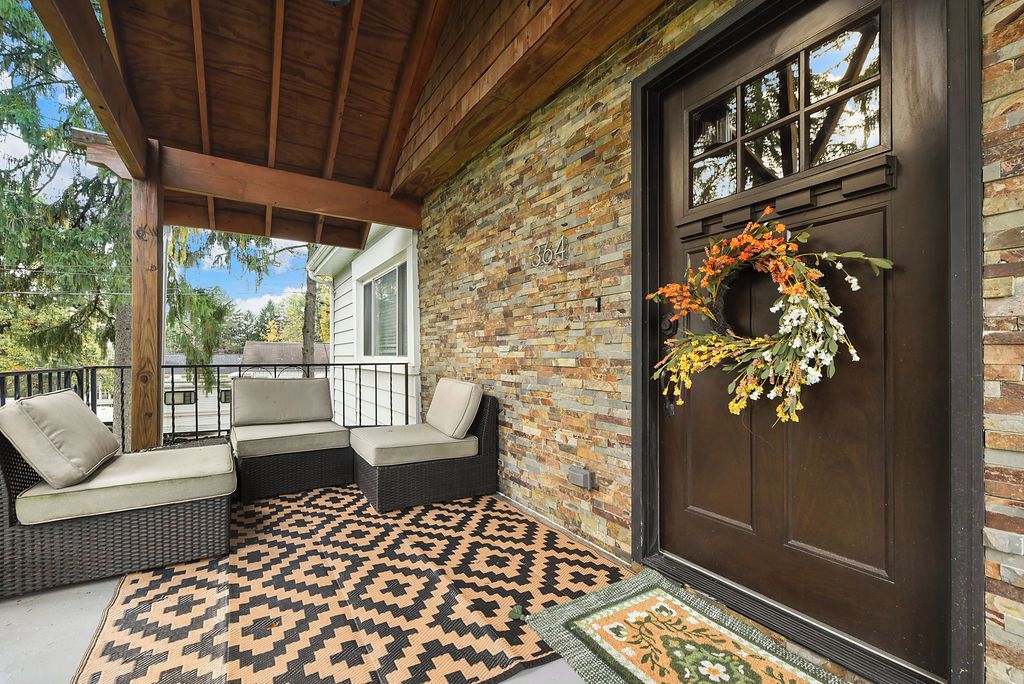 A covered porch with wicker furniture and a patterned rug, a brick wall, and a door with a wreath.