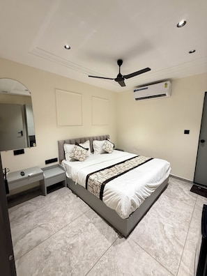 A modern bedroom with a bed, bedside tables, a mirror, an air conditioning unit, and a ceiling fan. - Hotel Pacific Comfort (Ahmedabad)