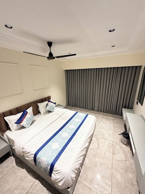 A hotel room with a bed, a ceiling fan, a window with curtains, and a desk with a chair. - Hotel Pacific Comfort (Ahmedabad)