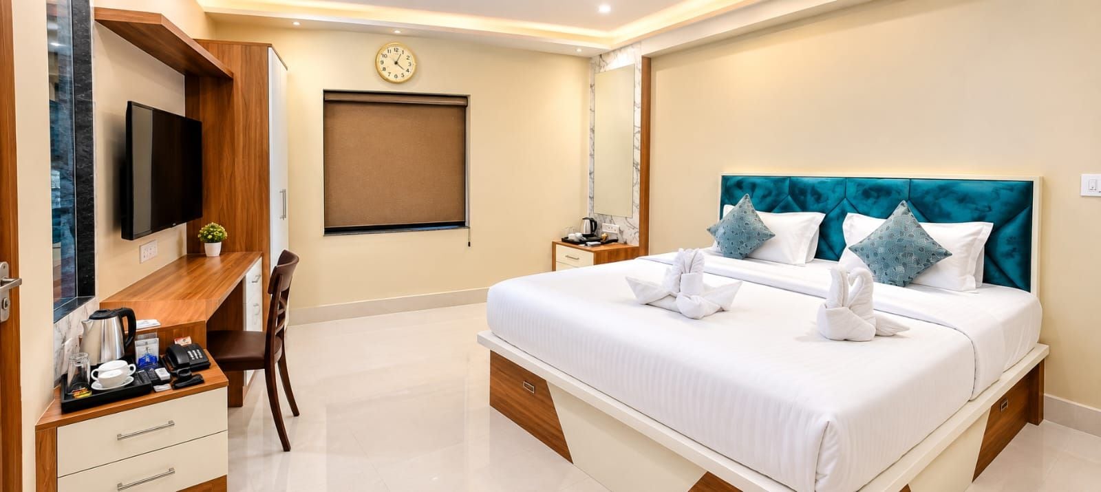 A hotel room with a bed, a desk with a flat-screen TV, a clock, and a window with a blind.