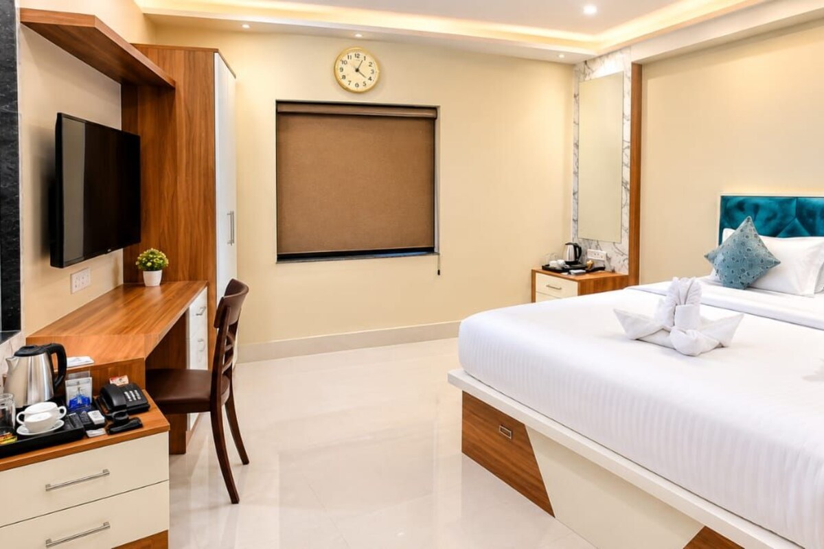 A hotel room with a bed, a desk with a flat-screen TV, a clock, and a window with a blind.