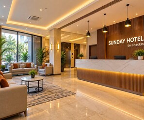Un espace d’accueil d’hôtel au design moderne, comprenant un comptoir d’accueil, un coin salon avec des canapés et une grande fenêtre offrant une vue sur l’extérieur.
