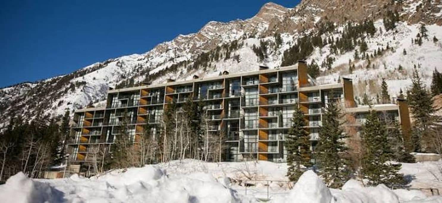 A multi-story hotel with a modern design, situated in a mountainous area with snow-covered peaks in the background.