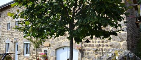 A stone building with a large tree in front, a white door, and a garden with flowers.