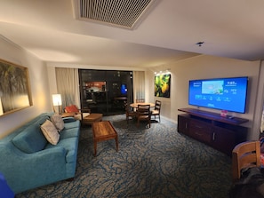 Ein Hotelzimmer mit einer Couch, einem Couchtisch, einem Fernseher und Blickrichtung Küche und Balkon.