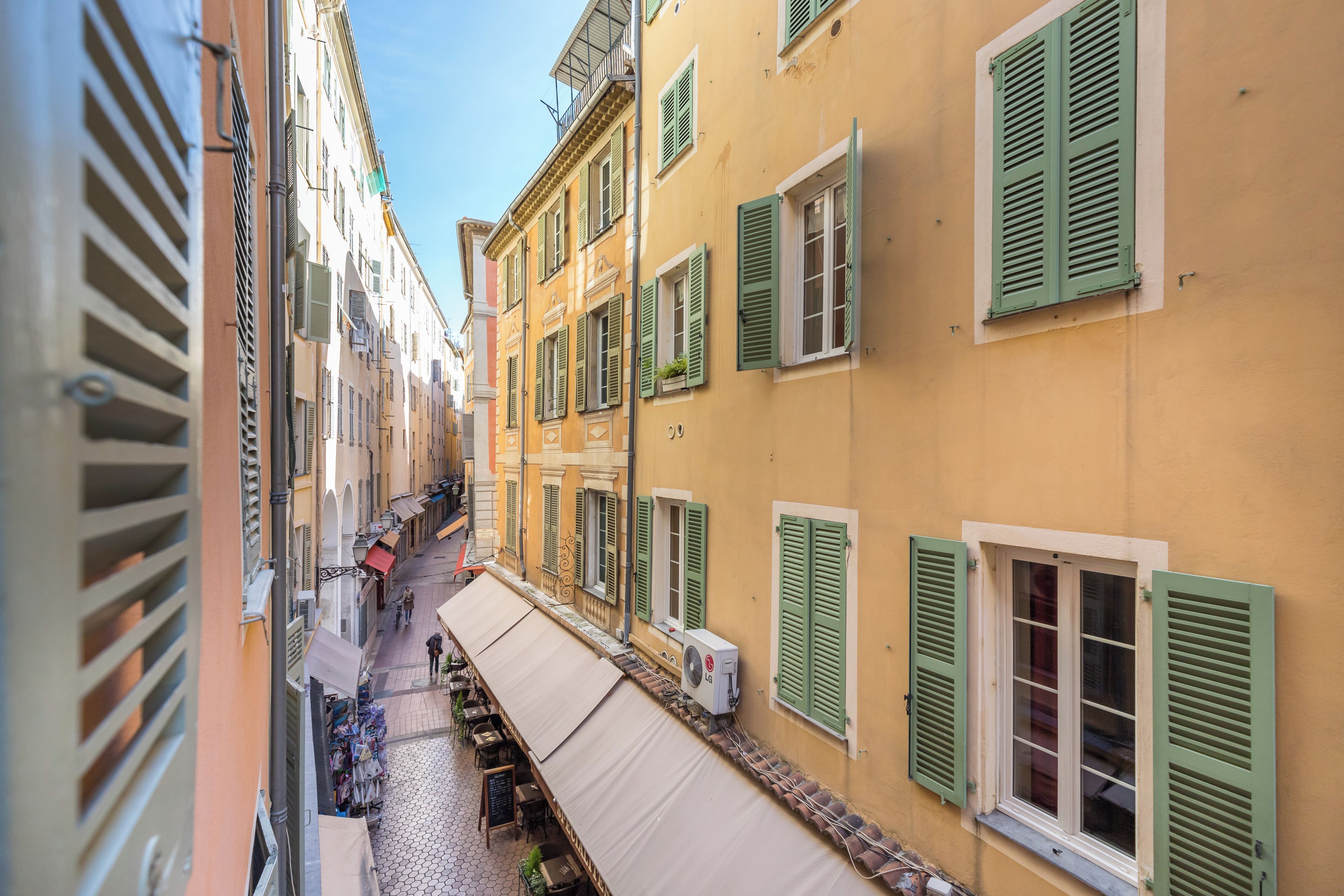 A narrow street with buildings on either side, featuring balconies and awnings.