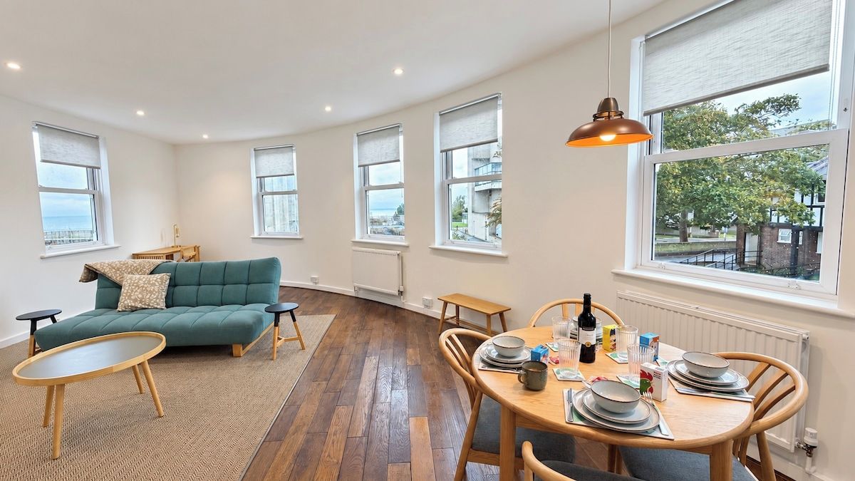 Amplia sala de estar con sofá, mesa de comedor para cuatro personas y ventanas grandes con vistas.