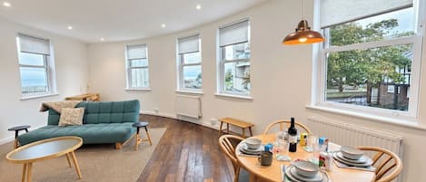 Amplia sala de estar con sofá, mesa de comedor para cuatro personas y ventanas grandes con vistas.