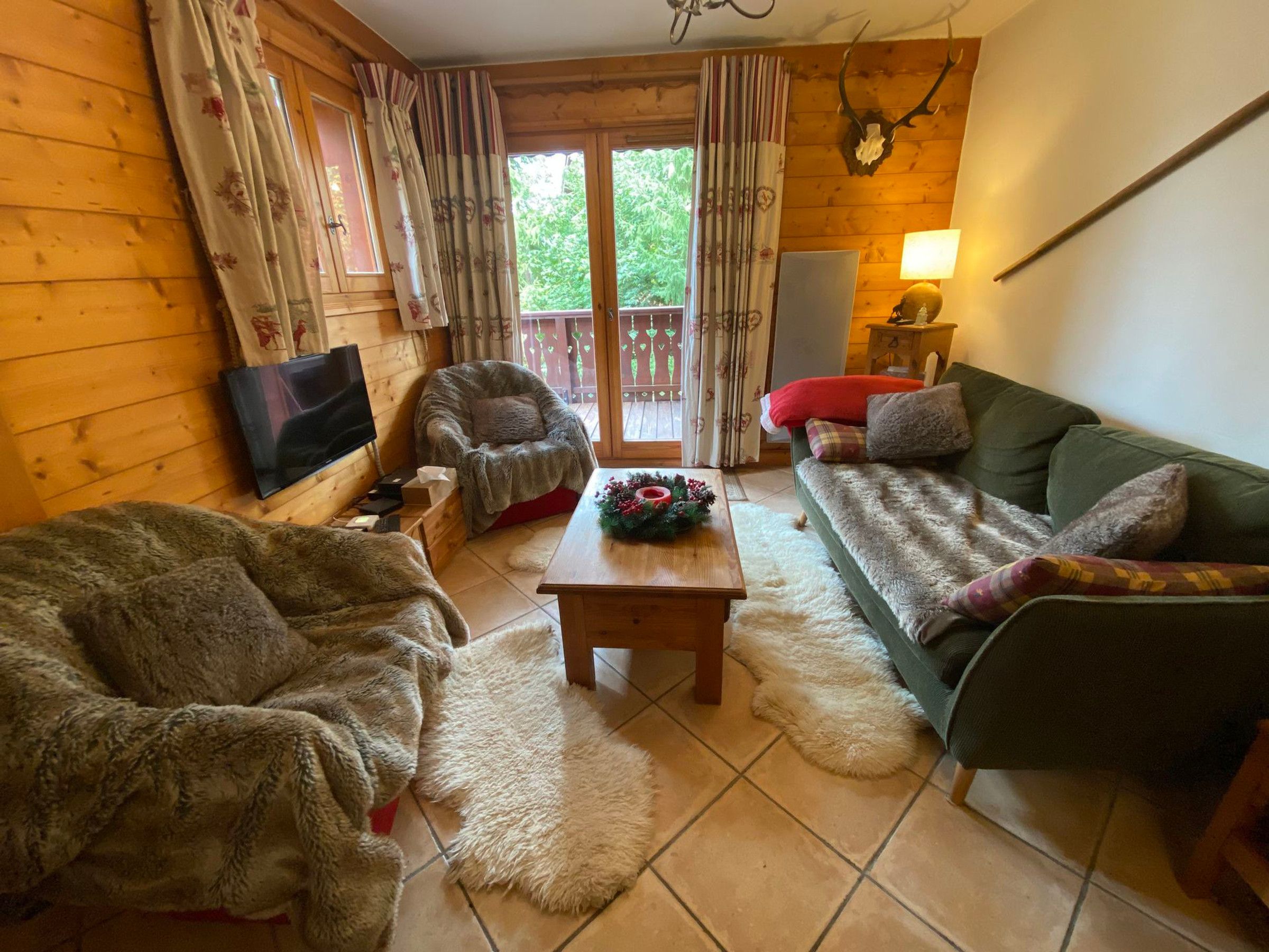 A cozy log cabin interior with a wooden floor, a green sofa, a plush armchair, a television, and a fireplace.