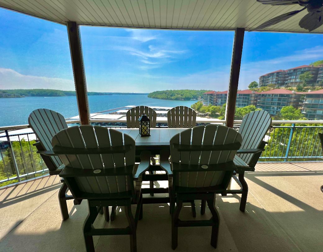 A covered outdoor seating area with a table and Adirondack chairs, overlooking a body of water and buildings.