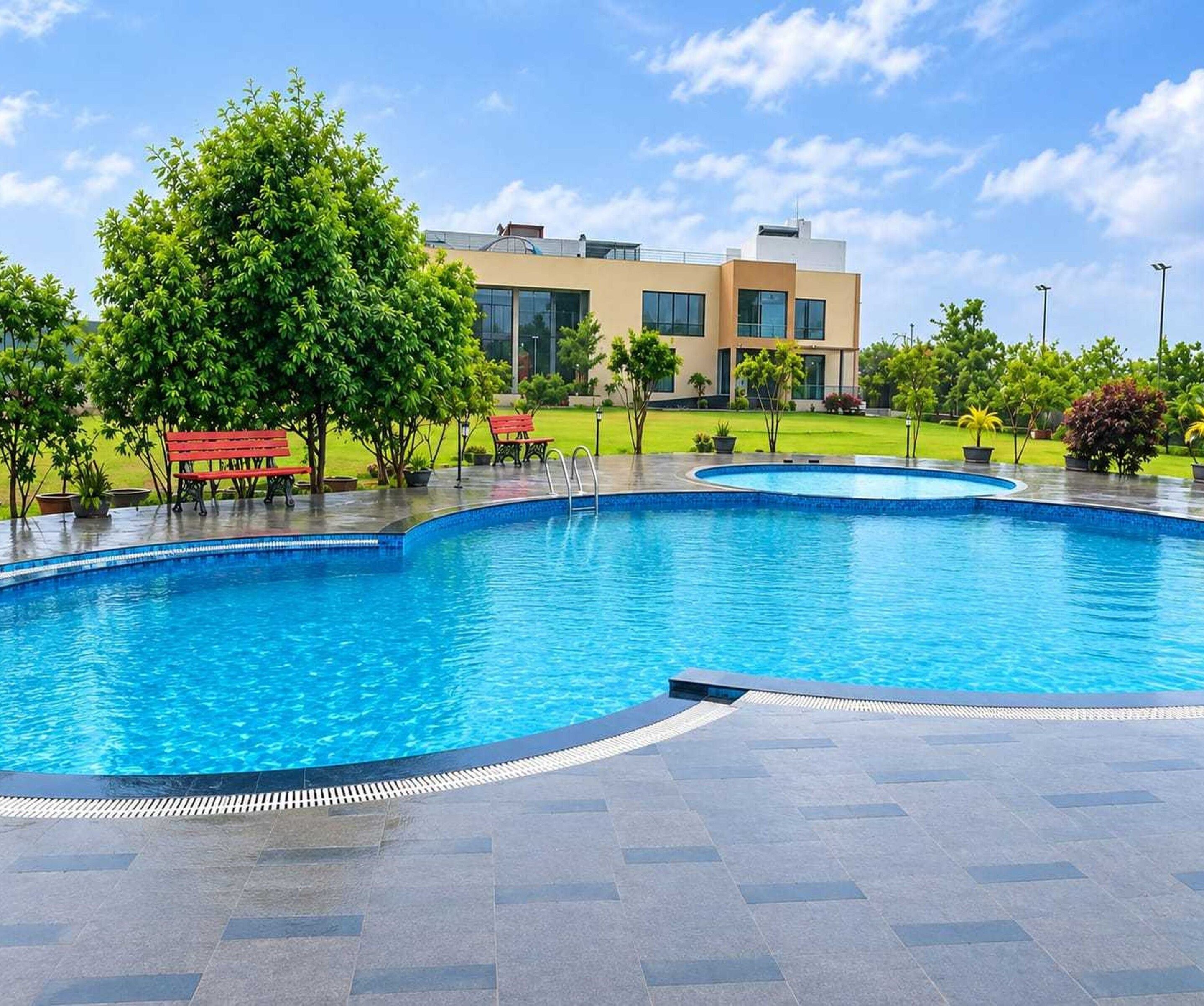 Une piscine à bord incurvé, entourée d’une terrasse dallée. On aperçoit des bancs et des arbres à l’arrière-plan.