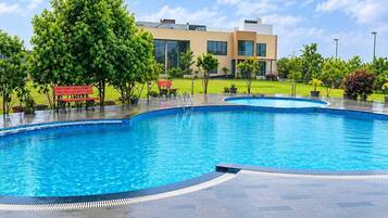 Une piscine à bord incurvé, entourée d’une terrasse dallée. On aperçoit des bancs et des arbres à l’arrière-plan.