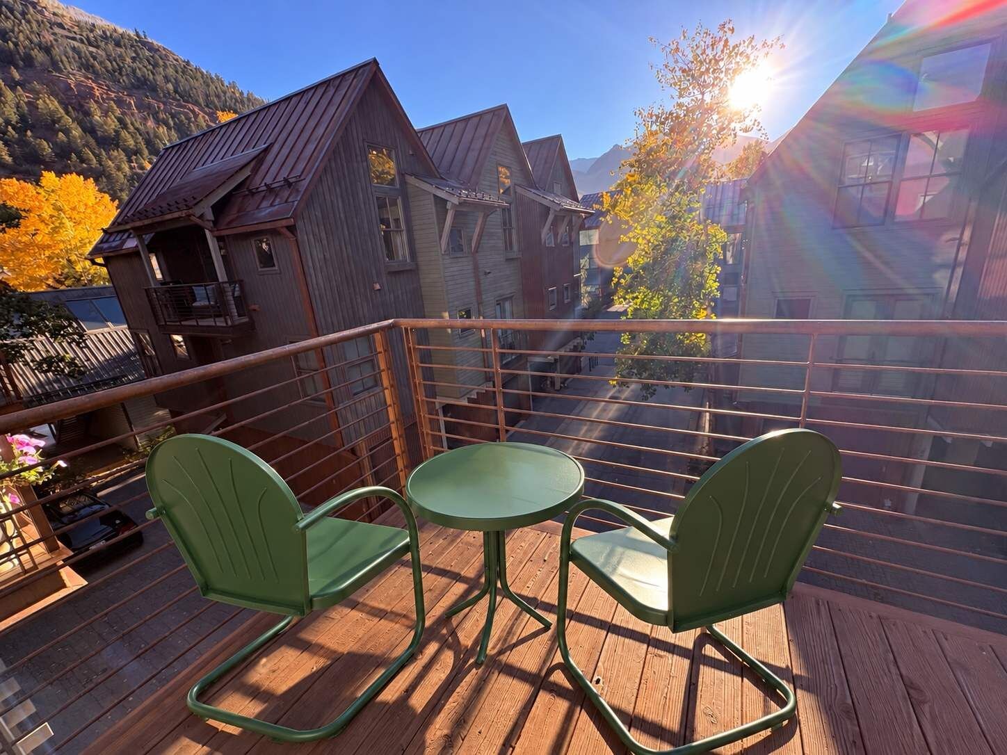 Una terraza de madera con dos sillas verdes y una mesa pequeña, con vistas a una calle con edificios y árboles otoñales.