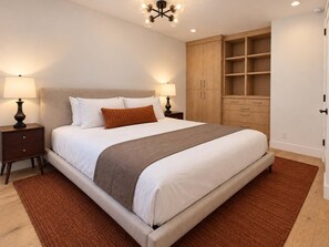 Una cama bien tendida con cabecera, mesitas de noche con lámparas y un armario de madera con estantes abiertos.