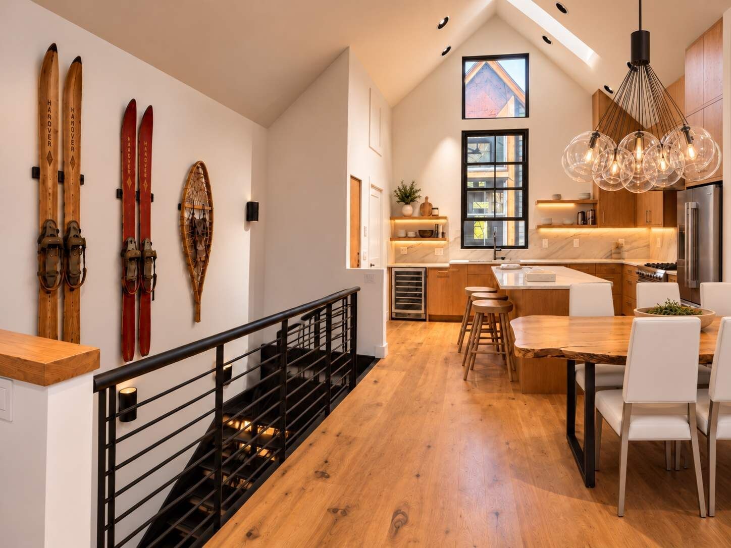 Una cocina moderna con piso de madera, un comedor con sillas blancas y una escalera con barandilla negra.