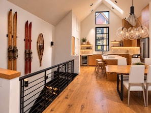 Cozinha moderna com piso de madeira, área de jantar com cadeiras brancas e escada com corrimão preto.