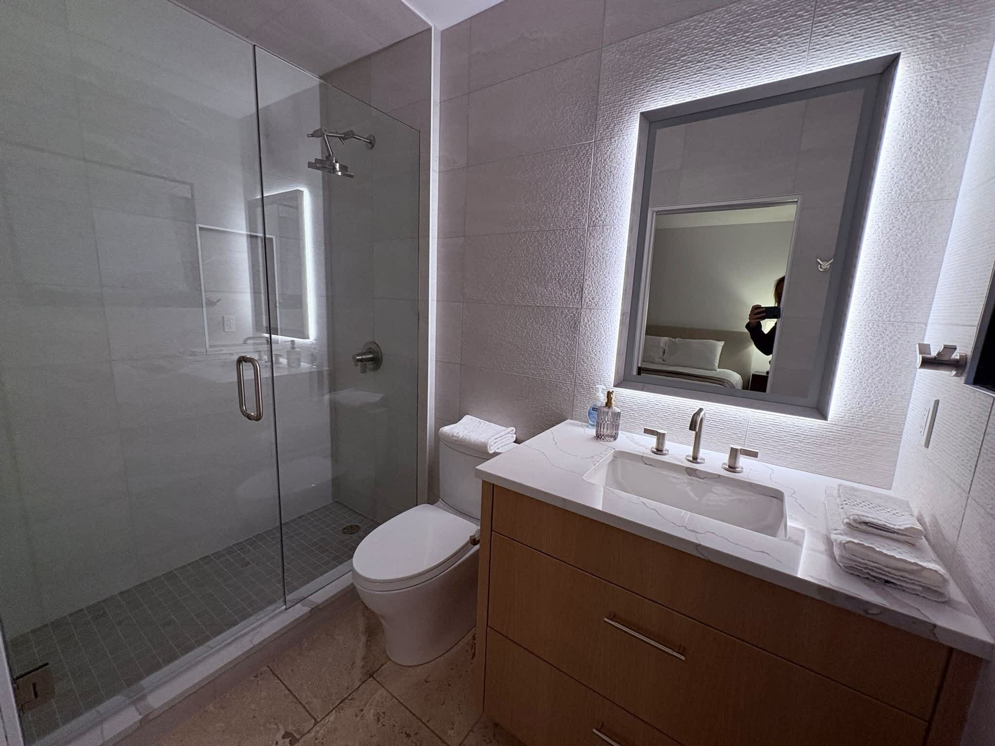 Un baño moderno con ducha de vidrio, inodoro, tocador con lavamanos y un espejo grande.
