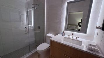 Un baño moderno con ducha de vidrio, inodoro, tocador con lavamanos y un espejo grande.