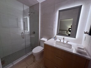 Un baño moderno con ducha de vidrio, inodoro, tocador con lavamanos y un espejo grande.