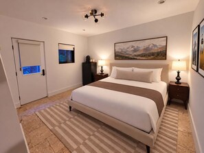 Una habitación de hotel con una cama grande, una mesita de noche con lámpara, un televisor montado en la pared y un cuadro de montañas.