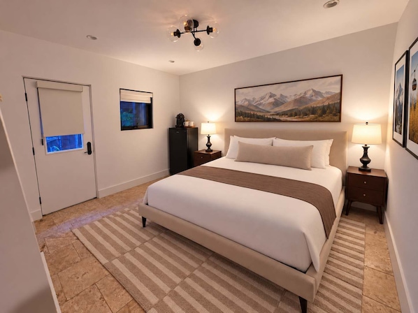 Una habitación de hotel con una cama grande, una mesita de noche con lámpara, un televisor montado en la pared y un cuadro de montañas.