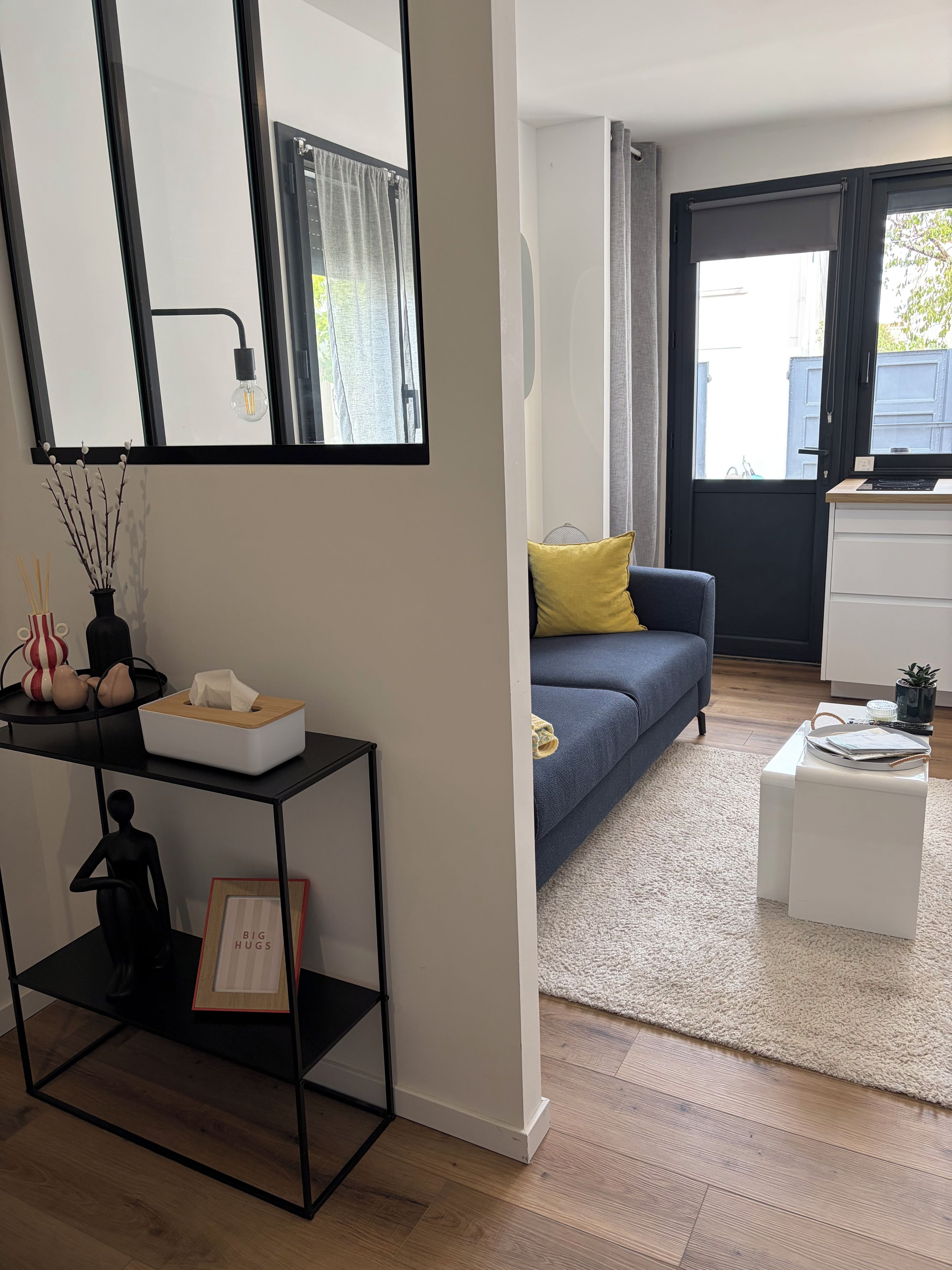 Een moderne woonkamer met een bank, een salontafel, een bijzettafeltje, een zwarte console met decoratieve objecten, een spiegel en een groot raam met gordijnen.