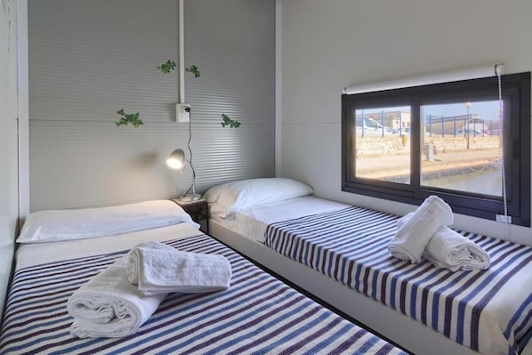 Habitación de hotel con dos camas individuales, cada una con ropa de cama y toallas a rayas. Se observa una ventana con vistas al exterior, y un aplique en la pared.