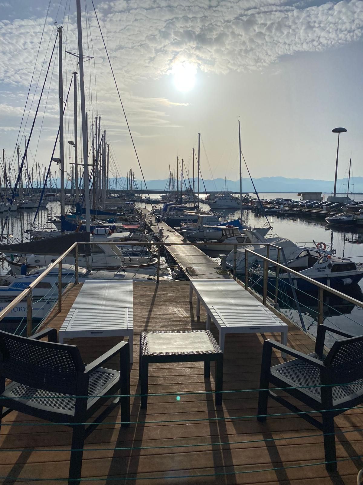 Ein Yachthafen mit mehreren vertäuten Booten, ein Holzdeck mit Bänken und ein klarer Himmel mit Sonnenschein.