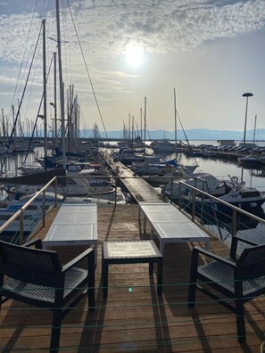 Un puerto deportivo con varias embarcaciones amarradas, una terraza de madera con bancos y un cielo despejado con el sol brillando.