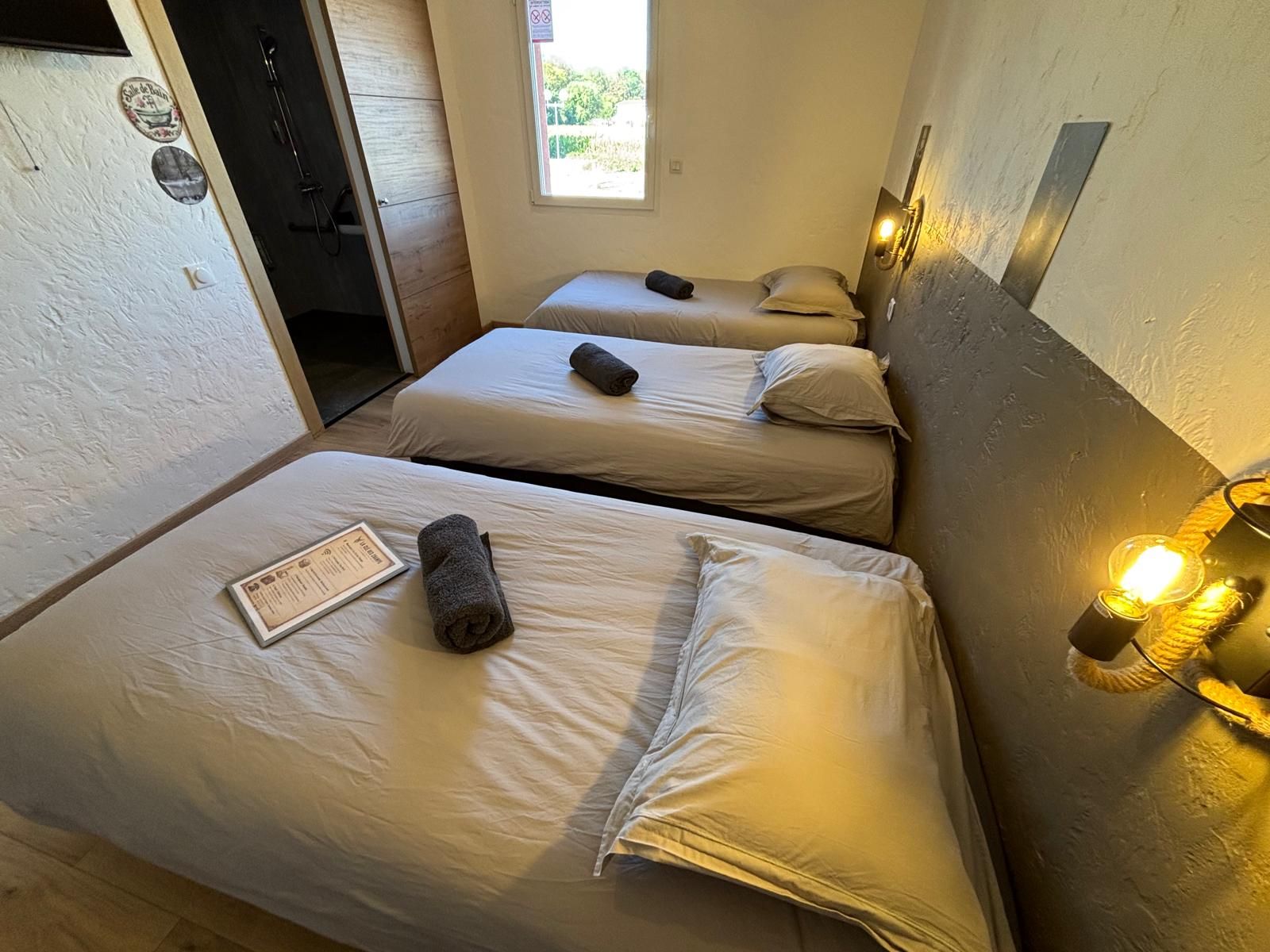 Une chambre d’hôtel avec deux lits simples, chacun équipé d’un oreiller et d’une serviette enroulée. La chambre dispose d’une fenêtre qui laisse entrer la lumière naturelle et d’un luminaire fixé au mur.