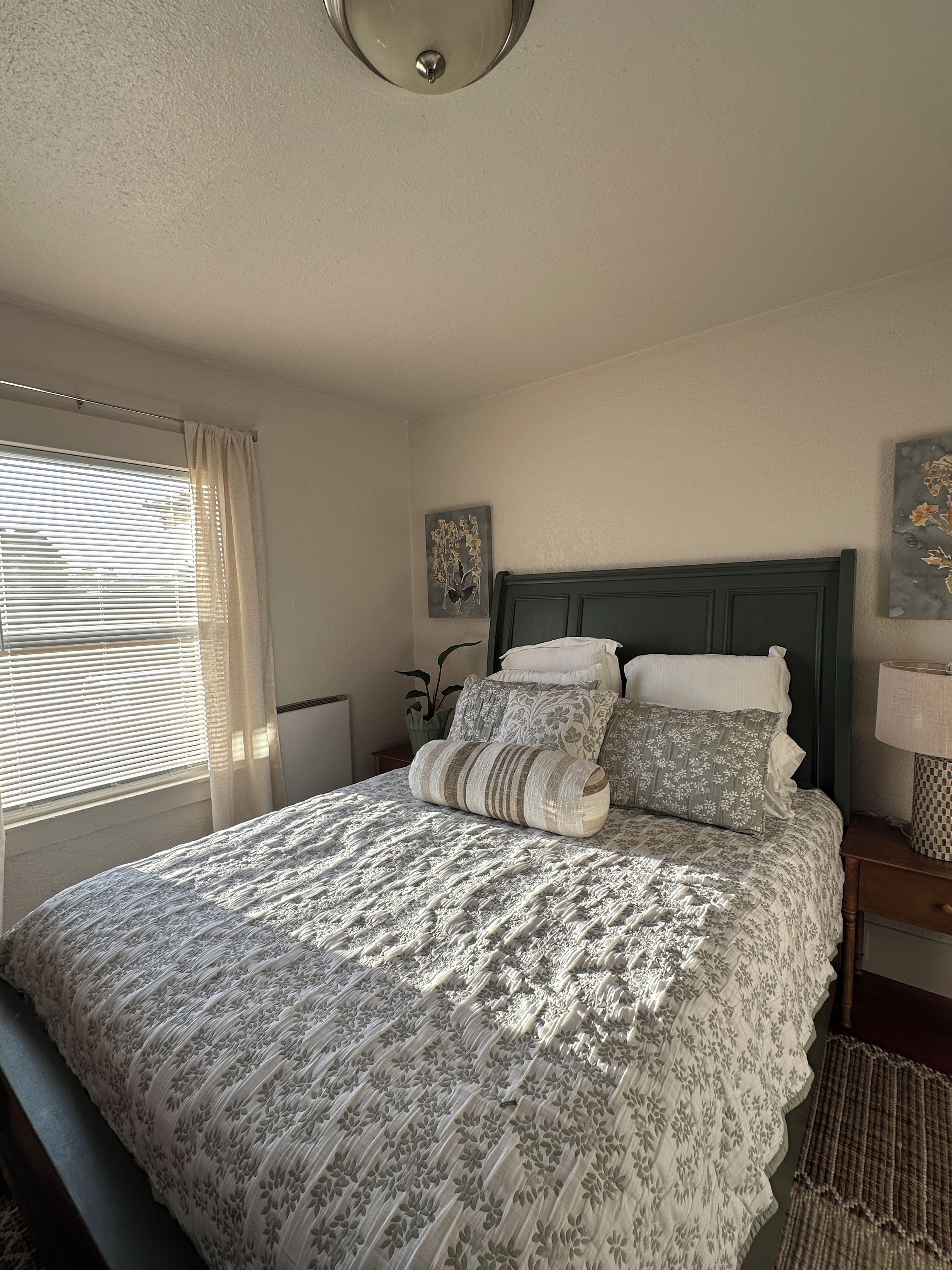 A bedroom with a bed, a nightstand, a lamp, a window with blinds, and a ceiling light.