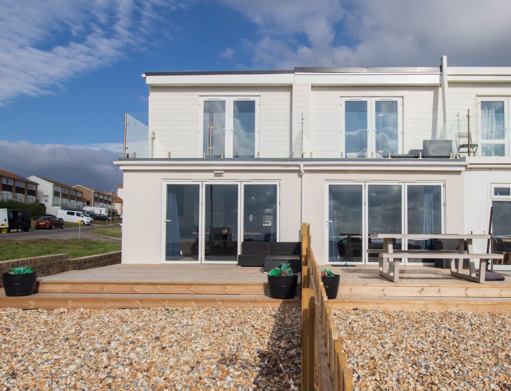 Un bâtiment moderne à deux étages doté de grandes portes coulissantes en verre, d’une terrasse en bois et d’un espace salon extérieur.