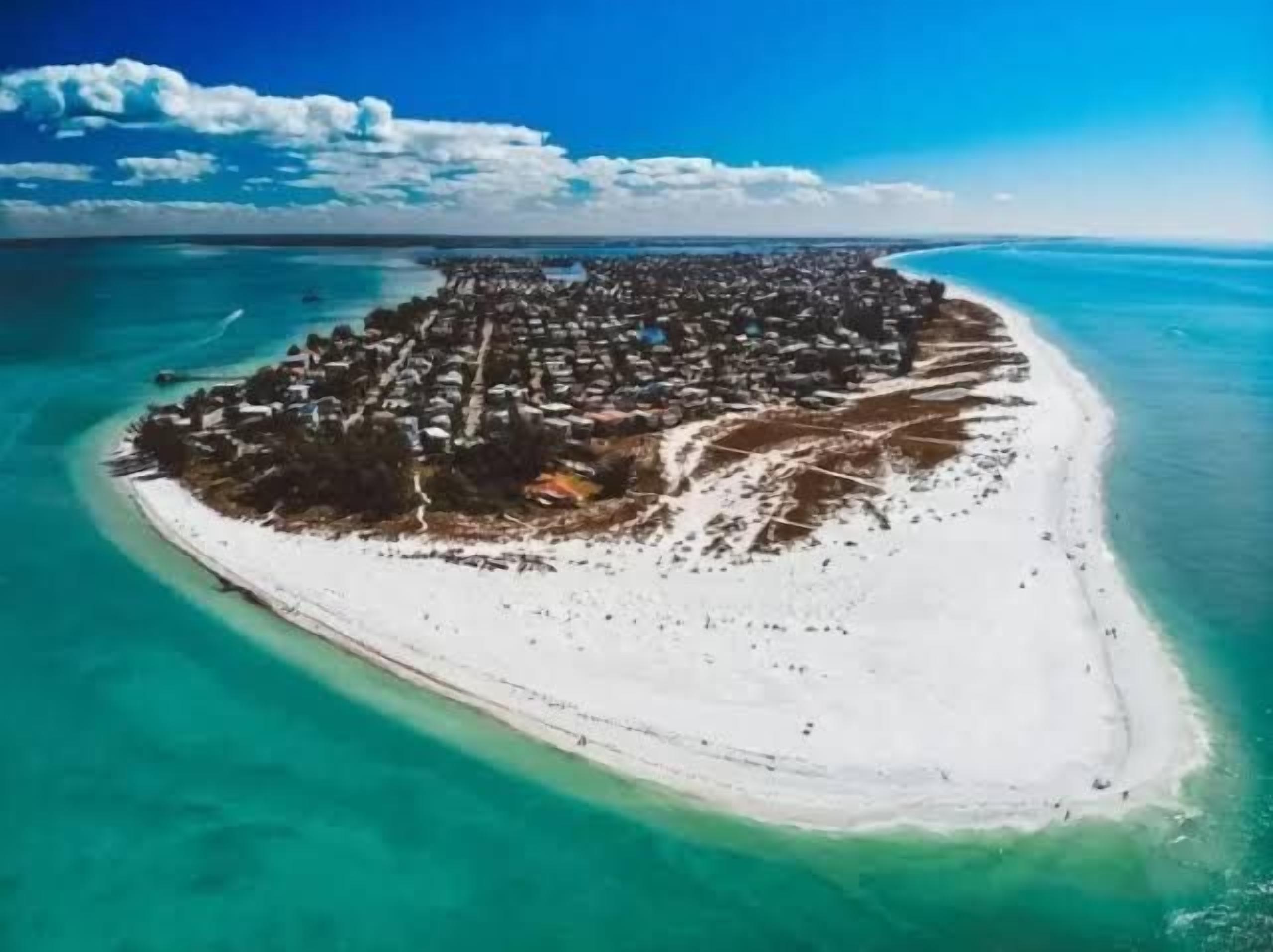 An aerial view of a coastal island with a dense settlement, clear turquoise waters, and a white sandy beach.