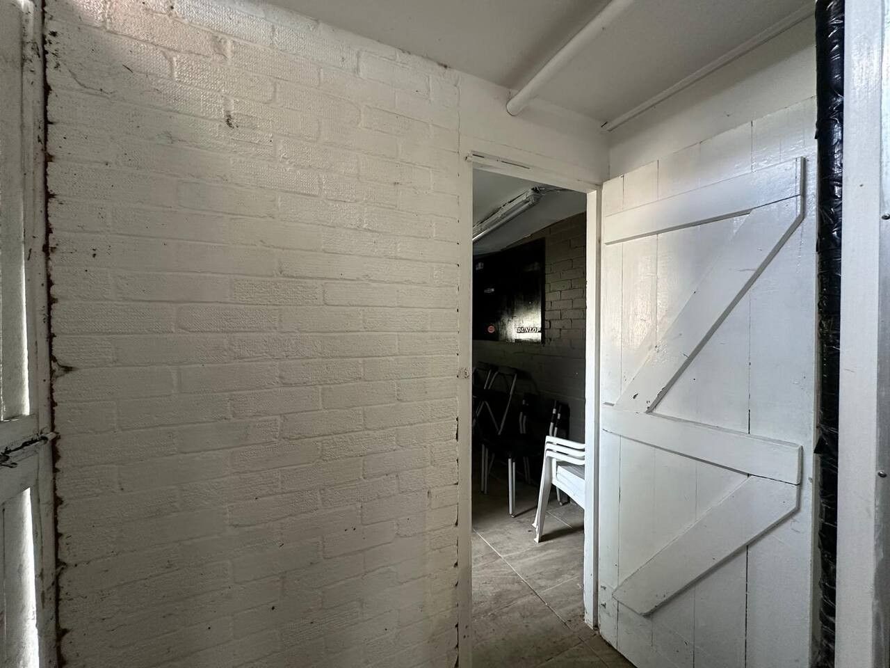 Een gang met witte bakstenen muren, een witte deur, en een gedeeltelijk zichtbare kamer met een stoel en een tafel.