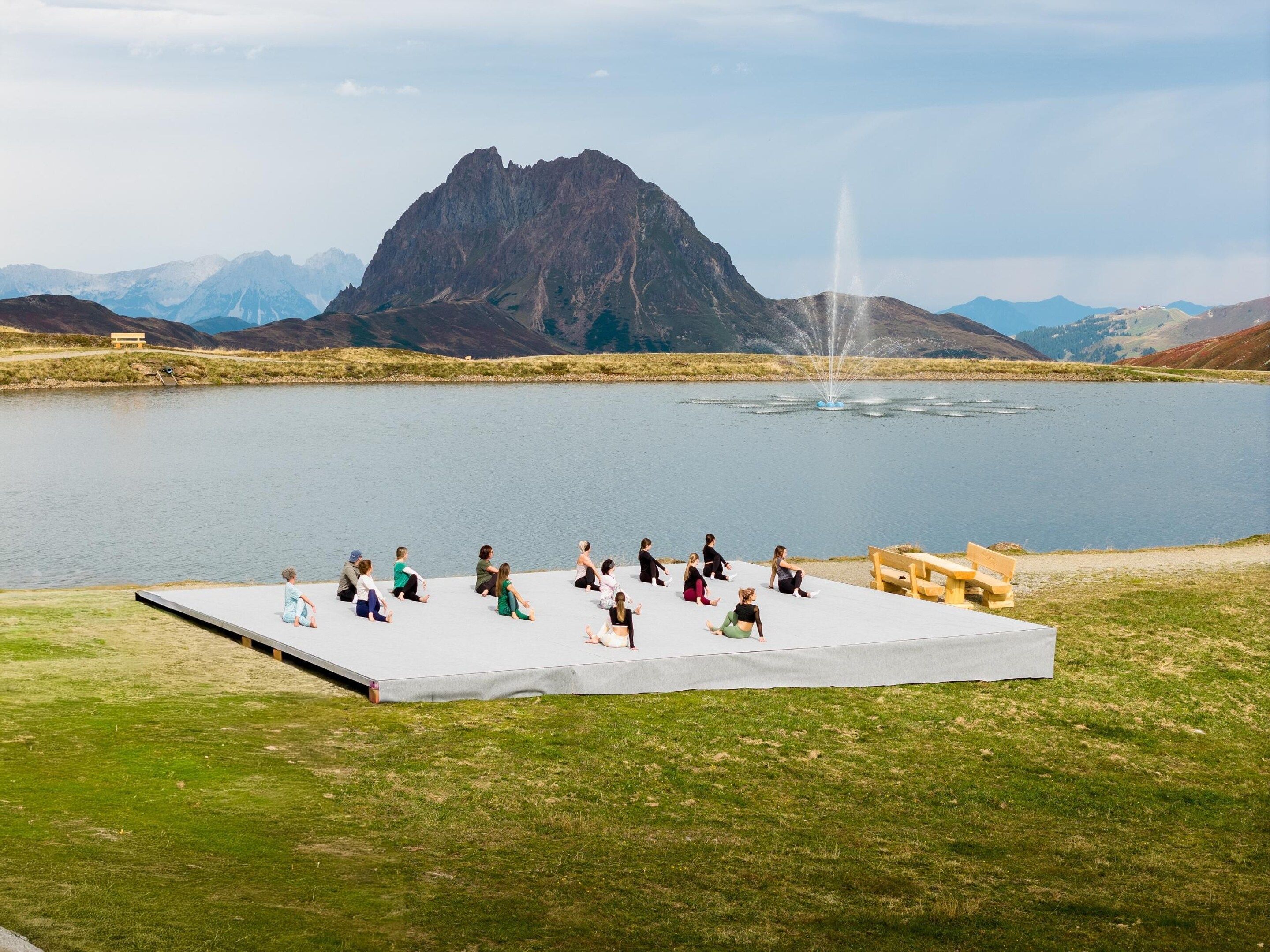 Un groupe de personnes est assis sur une scène, près d’un lac, avec des montagnes en arrière-plan.