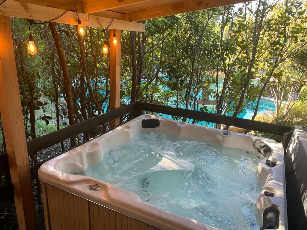 A hot tub with a wooden deck and a view of a swimming pool and trees.