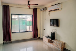Ein Zimmer mit Deckenventilator, Klimaanlage, an der Wand montiertem Fernseher und einem Fenster mit Metallgitter.