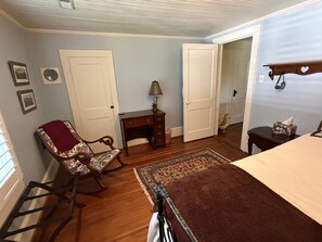 Een slaapkamer met een bed, een schommelstoel, een bureau en een deur naar een andere kamer.