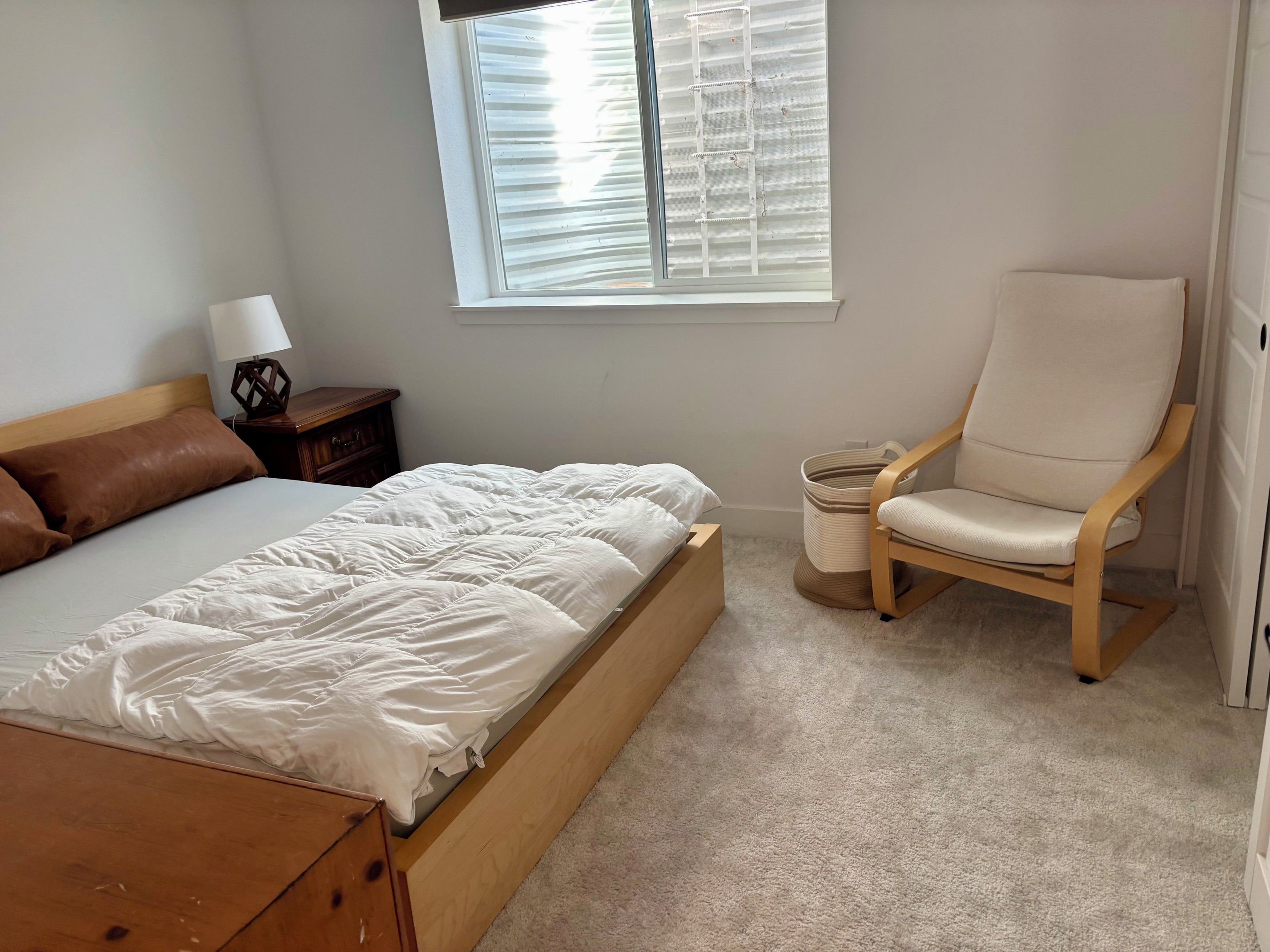 A bedroom with a bed, a chair, a nightstand, and a window with blinds.